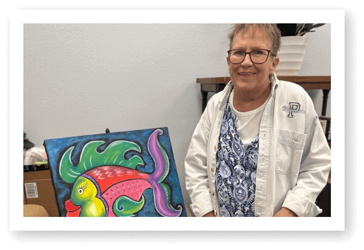 Elderly Woman Displaying Colorful Fish Painting During Art Class at Assisted Living Facility