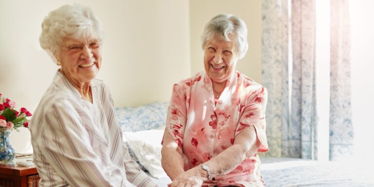 Two Elderly Women Sitting Together on a Bed in a Bright, Cheerful Memory Care Apartment